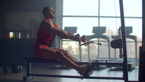 A-man-pulls-weight-to-his-stomach-sitting-in-the-simulator-performing-an-exercise-for-the-muscles-of-the-back-against-the-background-of-large-Windows-of-the-gym.-Powerful-male-athlete-doing-rowing-exercises